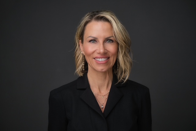 Woman with blonde hair wears a dark blazer and smiles at the camera.
